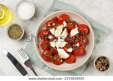 Roasted red tomatoes on branches with seasoned feta cheese on round off-white plate on kitchen napkins on grey background