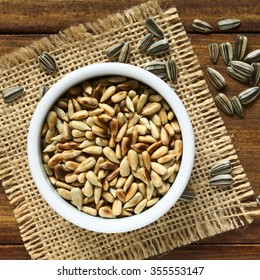 Roasted Sunflower Seeds In Small Bowl Surrounded By Whole Seeds With Hull, Photographed Overhead On Wood With Natural Light (Selective Focus, Focus On The Top Of The Roasted Seeds)