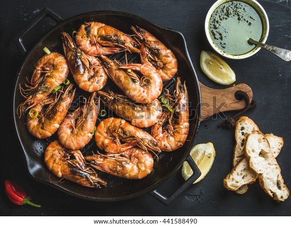 Roasted tiger\
prawns in iron grilling pan with fresh leek, lemon, bread and pesto\
sauce over black background, top\
view