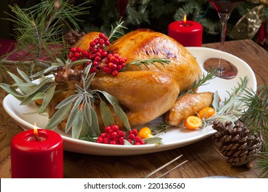 Roasted Turkey Garnished With Sage, Rosemary, And Red Berries In A Tray Prepared For Christmas Dinner. Holiday Table, Candles And Christmas Tree With Ornaments. 