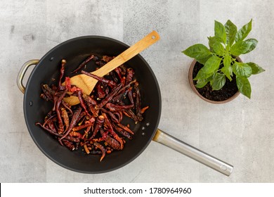 Roasting Dry Chili Peppers On Big Black Frying Pan. Little Chili Plant In Pot On Side. Wooden Spatula For Stirring. Gray Background, Top View.