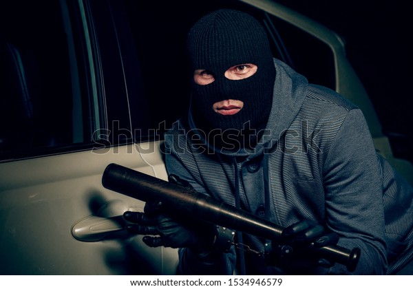 robber in a Balaclava tries to break the car glass\
with a baseball bat