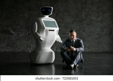 A Robot With Human Face And Body - Humanoid. Artificial Intelligence - AI. White Robot And Businessman In A Suit In A Modern Loft Office. Modern Robotic Technologies