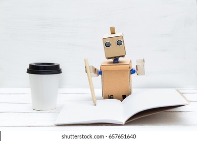 The Robot Is Working. He Holds A Pen In His Hand And Writes In A Notebook And Drinks Coffee