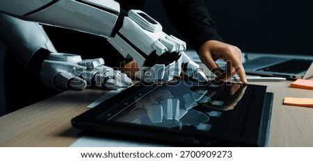 A robotic hand and a human hand work together on a sleek touchscreen device in a modern office, showcasing the future of technology and collaboration.Noogenesis