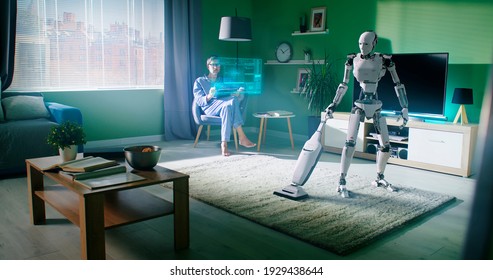 Robotic housekeeper using vacuum cleaner on soft carpet while tidying room near mature man browsing table with holographic interface at home