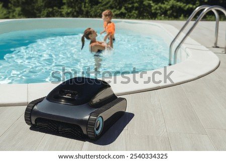 A robotic pool cleaner on the poolside with a mother and child enjoy swimming in clear blue water on background . Modern pool maintenance cleaning convenience. 