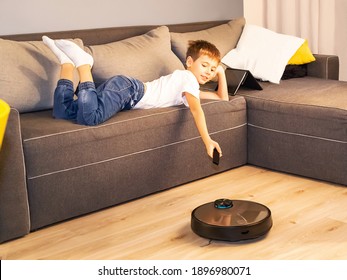 Robotic Vacuum Cleaner Clean Floor, While Boy Playing With Tablet Computer On The Sofa. Child Easily Controlling Vacuum Cleaner With Remote Control. Smart Home, Housework, Cleaning Technology Concept.