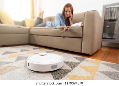 Robotic vacuum cleaner cleaning the room while woman relaxing on sofa. Woman controlling vacuum with remote control.