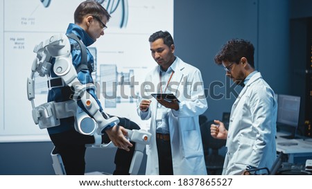 In Robotics Development Laboratory: Engineers and Scientists Work on a Bionics Exoskeleton Prototype with Person Testing it. Designing Wearable Exosuit to Help Disabled People, Warehouse Workers