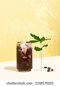 Robusta Iced Coffee Drink, Coffee Beans And Leaves On A Yellow Background. Can-shaped Glass Cup Of Coffee With Non-dairy Milk. Reusable Drinking Straw. Concept Of Sustainable Food And Drinks.