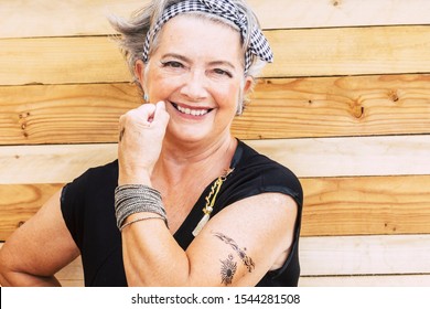 Rock And Alternative Cheerful Senior Caucasian Woman Show Her Tatoo On The Arm Smiilng At The Camera - No Limit Age To Have Fun And Enjoy Lifestyle For Old People - Wooden Background