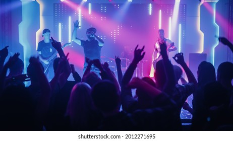 Rock Band with Guitarists and Drummer Performing at a Concert in a Night Club. Front Row Crowd is Partying. Silhouettes of Fans Raise Hands in Front of Bright Colorful Strobing Lights on Stage. - Powered by Shutterstock