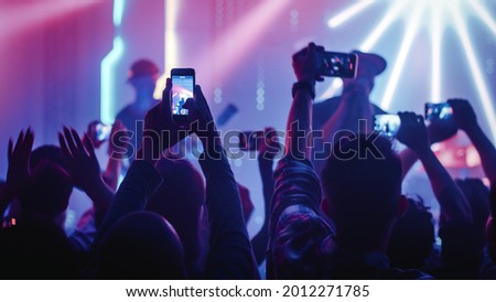 Rock Band with Guitarists and Drummer Performing at a Concert in a Night Club. Front Row Crowd is Record Video on Their Mobile Phones. Party in Front of Bright Colorful Strobing Lights on Stage.
