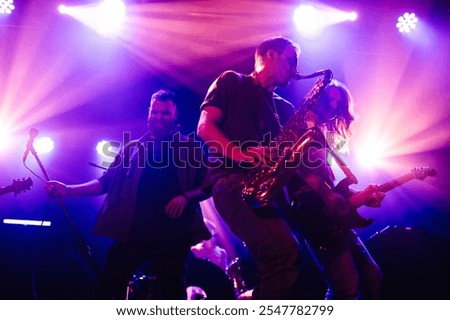 Rock band performs on stage with saxophone, guitar, and vocals under purple lights, captivating the audience with their energy and passion for music