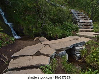 Rock bridge on hiking trail of stone material constructed by Nepali Sherpas leading to famous peak of mountain Reinebringen, Moskenesøya island, Lofoten, Norway with small waterfall and green plants.