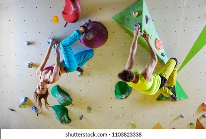 Rock climbers in bouldering gym climbing up overhanging wall. Young woman hanging upside down and smiling, male climber looking at her.