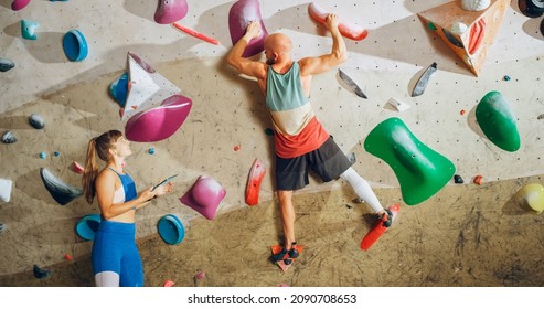 Rock Climbing Instructor Giving Lesson to a Beginner on a Bouldering Wall in a Gym. Female Athlete Holding Tablet Computer and Showing Correct Climbing Techniques to Strong Fit Male. - Powered by Shutterstock