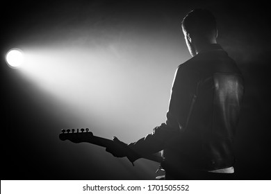 Rock Guitarist Man In Leather Jacket Standing His Back In Smoky Studio Or Stage Masterfully Playing Electric Guitar. View Of Unrecognizable Musician In The Spotlight.