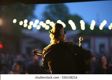 Rock musician with a guitar and a hat on a stage on blurred background of street concert audience