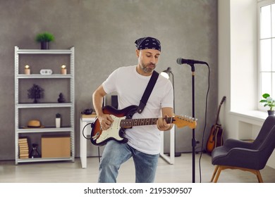 Rock musician playing music in home studio. Guitarist rehearsing new song. Bearded middle aged man in bandana leisurely strumming on electric or bass guitar standing in rehearsal room with