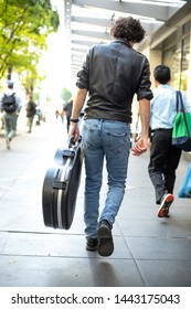 Rock Musician Wearing A Leather Jacket And Carrying A Guitar Case On A City Sidewalk, In A Music Busker Concept