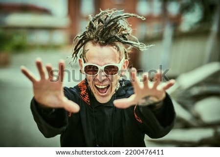 Rock and punk culture. An extraordinary mature man with dreadlocks on his head and tattoos on his body stretches his arms forward and shouts with joy, standing on a city street.