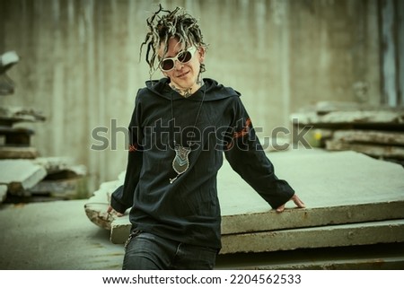 Rock and punk culture. An extraordinary mature man with dreadlocks and tattoos wearing black clothes with ethnic ornaments stands on the backstreet and smiles.