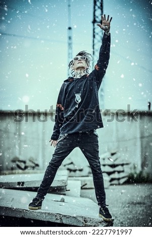 Rock and punk culture. Extraordinary mature man with dreadlocks and tattoos in black clothes with ethnic ornaments stands on the backstreet under snowfall looking up at the sky with his hand raised.