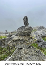 A rock stack stands tall on a mountain with fog covering the landscape. Green grass and various stones surround the stack, adding to the natural setting on a cloudy day.