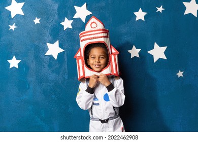 Rocket afro-american little boy in space suit playing astronaut on blue background with white hand-made stars. Happy mixed race child with handcrafted spacecraft. Childhood, creative and imagination.