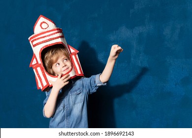 Rocket boy kid with hand-made toy spaceship on head playing in astronaut. Future spaceman pointing on blank space on background for advertising text. Space child game and education concept.