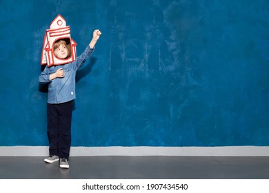 Rocket little caucasian boy kid with toy spacecraft on head showing cosmic flight with arm raised up. Cute child playing in astronaut posing on blue background with copyspace for advertising.