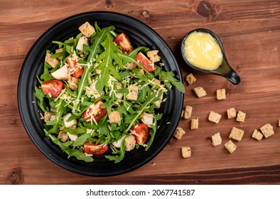 Rocket salad with tomato, crouton, herb roasted chicken, parmesan cheese in black dish over sackcloth together with a sauce cup full of mayonnaise and extra crouton  on the side dish on wooded table.