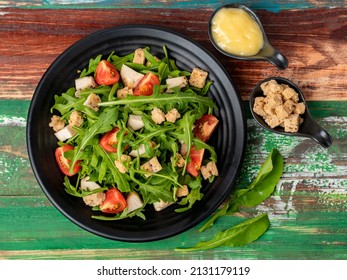 Rocket salad with tomato, crouton, herb roasted chicken in black dish together with two tiny sauce cups full of mayonnaise and extra crouton on colourful wooden table.