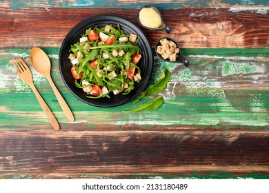 Rocket salad with tomato, crouton, herb roasted chicken in black dish together with wooden utensil and two tiny sauce cups full of mayonnaise and extra crouton on colourful wooden table