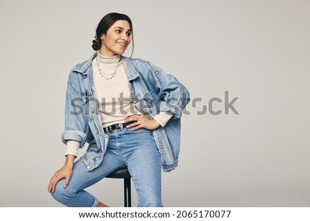 Rocking denim wear. Fashionable young woman sitting on a chair against a grey background. Happy young woman looking away with a smile on her face in a modern studio.