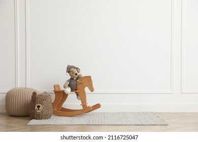 Rocking horse with bear toy, pouf and wicker basket near white wall in child room, space for text. Interior design