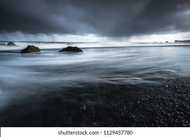 Rocks On A Black Sand Beach In Iceland With Reflection In The Sea And A Dark Sky In Misty Moody Weather With Dark Colors And A Rough Sea In Landscape Format With A Storm Approaching