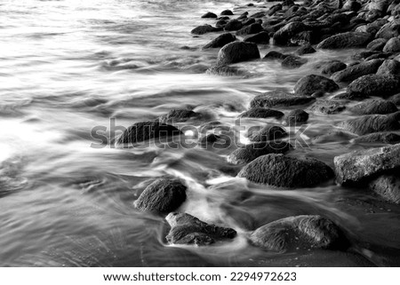 Rocks and pebbles on a beach in the Caribbean sea. Surf water motion with longtime exposure showing flowing streams and rivulets. Black and white greyscale with low contrasting evening sun light.