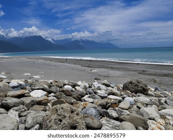 A rocky beach with smooth stones, clear turquoise water, and a backdrop of mountains under a partly cloudy sky, creating a serene and picturesque coastal scene. - Powered by Shutterstock