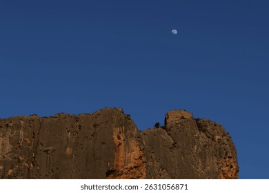 A rocky cliff with ancient ruins on top stands under a clear blue sky, with the moon visible above. The scene is captured through a window, showing a faint reflection of the photographer. - Powered by Shutterstock - Get 15% off with code: PIKWIZARD15