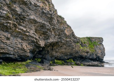 A rocky cliff with a beach in the background. The cliff is covered in moss and has a greenish tint. The beach is sandy and has a few rocks scattered around - Powered by Shutterstock