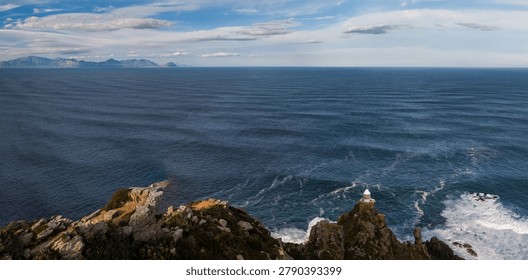 Rocky cliffs drop into a deep sea as waves churn below a white lighthouse at Cape of Good Hope, Cape Town. Distant mountains line the horizon under scattered clouds in daytime.
