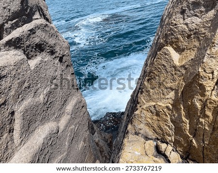Rocky cliffs frame the ocean as waves crash between them, showcasing a coastal landscape under clear blue skies and bright sunlight