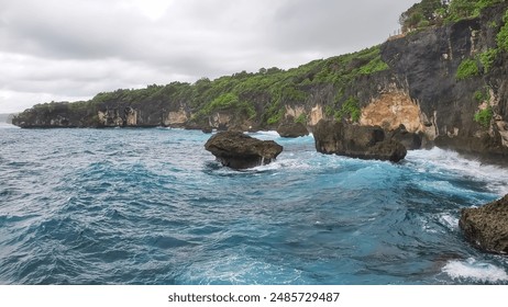 rocky cliffside with lush greenery overlooking a turquoise ocean with crashing waves. The water is a beautiful shade of blue, and the sky is a soft gray. - Powered by Shutterstock