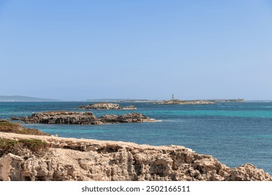 The rocky coast of Ibiza stretches towards the turquoise waters, with distant lighthouses of Formentera visible on the horizon, framed by rugged cliffs and clear blue skies - Powered by Shutterstock