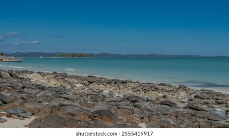 The rocky coast of the ocean. Boulders are scattered near the turquoise water. Boats at sea. There is a green tropical island in the distance. Mountains on the horizon. Clear  sky. Madagascar.  - Powered by Shutterstock