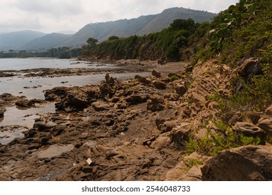 A rocky coastal path lined with lush greenery, overlooking the sea and distant hills. The rugged landscape provides a serene and natural environment by the shore - Powered by Shutterstock