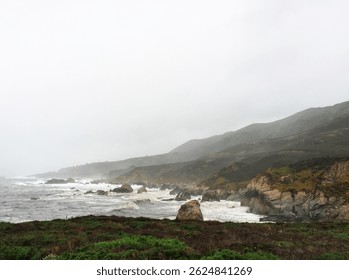 Rocky coastline with crashing waves and misty hills under overcast sky
